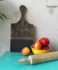Belles and Whistles When Life Gives You Lemons - Stencil.  A wooden cutting board that is chalk painted black on the lower half and has the When Life Gives You Lemons stencil applied to the top and handle with black chalk paint.  A yellow and red bowl sits on the table holding red applies; an apple is sitting on the table in front of the cutting board; and a brown wooden rolling pin is sitting in front of the bowl.  All sits on a blue table, ion front of a gray wall.  Greenery sits at the left of the table.