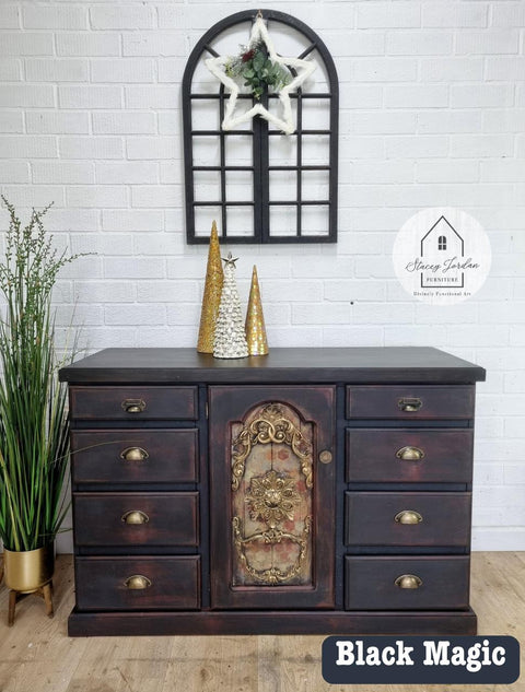 A dark wooden dresser with intricate floral design on the center door, adorned with golden decorations and small trees. It sits against a white brick wall, under a black arched mirror. A plant in a gold pot is on the left side.