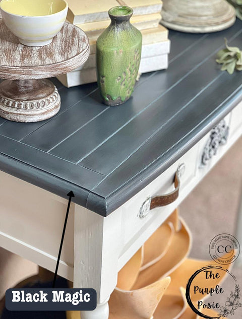 A rustic table with a stained black top labeled "Black Magic." The table has a white lower half with a brown leather handle, decorative vases, and stacked dishes on top. A logo in the corner reads "The Purple Posie.