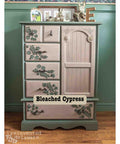 Wooden dresser with floral designs and 'Bleached Cypress' label against a green wall.