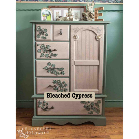 Wooden dresser with floral designs and 'Bleached Cypress' label against a green wall.