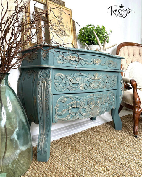Elegant vintage-style teal dresser with ornate carvings, situated on a woven rug. It holds a framed artwork, potted plant, and a wooden armchair. A glass vase with branches stands nearby, adding rustic charm to the scene.
