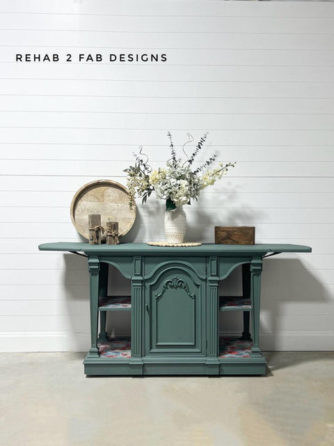 A green vintage sideboard against a white shiplap wall, featuring floral-patterned shelves. It is adorned with white flowers in a vase, a round tray, and a small wooden box. Text on the wall reads "Rehab 2 Fab Designs.