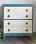 A vintage-style three-drawer chest with teal sides and white fronts featuring ornate brass knobs. The chest stands on curved legs, surrounded by a decorative vase and green potted plants. "McKay Designs" is in the bottom corner.
