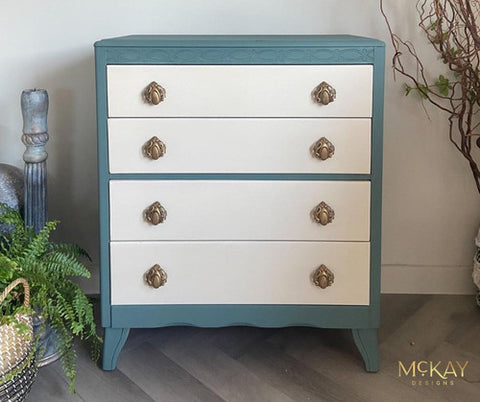 A vintage-style three-drawer chest with teal sides and white fronts featuring ornate brass knobs. The chest stands on curved legs, surrounded by a decorative vase and green potted plants. "McKay Designs" is in the bottom corner.