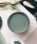 A flat lay of a green-themed arrangement featuring a jar of sage green paint, various shades of green fabric, a green purse, and sprigs of dried foliage on a white surface.
