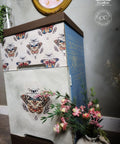 A two-drawer chest with butterfly designs on the front and floral patterns on the sides stands on a wooden floor. A vase with pink flowers is placed at its base. A round, gold-framed mirror hangs on the wall above.