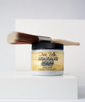 A paintbrush rests on top of a container of Dixie Belle Bonding Boss primer in gray, placed on a white geometric surface with a minimalist background.