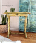 A vintage yellow entryway table with the Bone Inlay Stencil applied in decorative patterns with a crystal drop drawer pull, set against a pale blue rustic shutter backdrop, adorned with a small green plant on top, and complemented by indoor plants and a cozy white fur rug