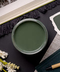 A top view of an open jar of dark sage green paint surrounded by fabric samples, a book, and white flowers on a dark surface. The arrangement suggests a color-coordinated design theme.