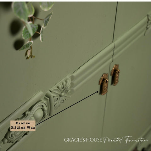 Close-up of a sage green cabinet with ornate detailing and bronze handles. A label pointing to the detail reads "Bronze Gilding Wax." Eucalyptus leaves hang on the left. Text at the bottom says "GRACIE'S HOUSE Painted Furniture.