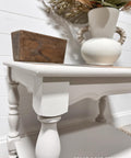 A white wooden side table with turned legs holds a rustic wooden box and a white vase with handles filled with dried foliage. The background features white shiplap walls. "REHAB 2 FAB DESIGNS" is written at the bottom.