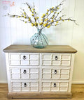 A white and wood dresser with six drawers stands against a white paneled wall. On top, there is a large clear glass vase holding long branches with yellow blossoms. The floor is light wood.