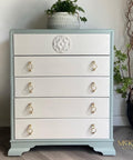 A small, five-drawer dresser with light blue edges and white drawers, featuring gold ring pulls. A white decorative medallion is on the top drawer. A potted plant sits on top, with another plant in a black basket nearby.