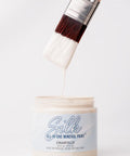 A hand holds a paintbrush dripping with white paint above an open container labeled "Silk All-In-One Mineral Paint, Chantilly" against a plain white background.