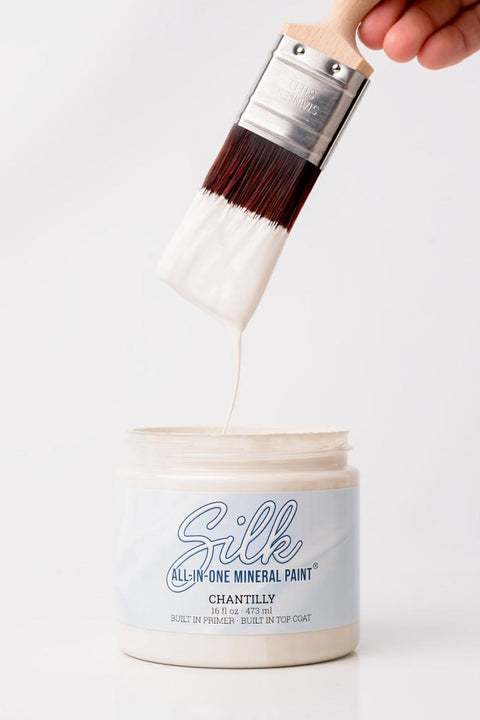 A hand holds a paintbrush dripping with white paint above an open container labeled "Silk All-In-One Mineral Paint, Chantilly" against a plain white background.