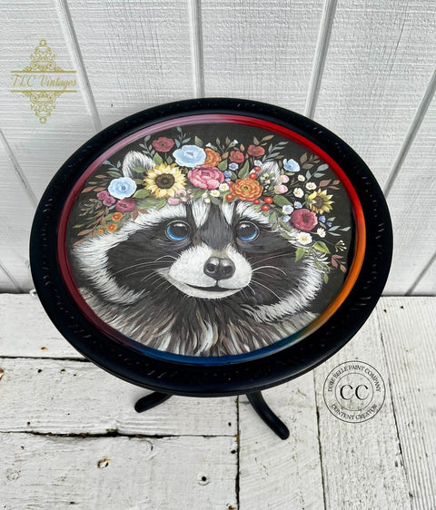 A round wooden table features a detailed painting of a raccoon wearing a colorful floral crown. The table's top has a black rim, and the legs are black as well. The background behind the table is white paneling, and logos are visible in the background.