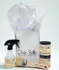 A set of Dixie Belle paint products, including a spray bottle, one small jar of Caviar paint, a paint brush, and a jar of clear coat, displayed in front of an open box with tissue paper.
