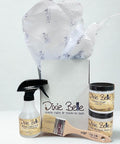 A gift box with tissue paper contains Dixie Belle products: a spray bottle, a paintbrush, and two small jars labeled "Clear Coat Satin" and "Chalk Mineral Paint Fluff", all against a plain white background.