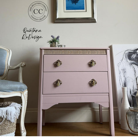 A small pink dresser with ornate handles is placed against a white wall. To the left, a light blue cushioned chair with white wooden frame is visible. Art pieces, plants, and decorative items are arranged around the scene.