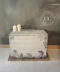 An off-white painted wooden chest with intricate black illustrations from the Cottage Toile Transfer, placed against a gray wall. The chest has two lit small white candles and a small branch on top. A jute mat lies underneath it. A logo for "Dixie Belle Paint Company Content Creator" is seen on the picture.