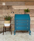 Dixie Belle Paint Antebellum Blue Chalk Mineral Paint is the perfect paint for any DIY project! Classic Drop Lid Desk painted with Antebellum Blue Chalk Mineral Paint. Accented with gold hardware and staged with chair and planter against hardwood wall.