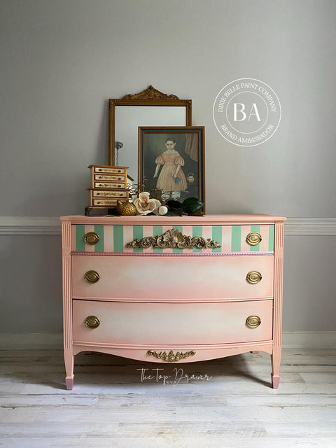 Dixie Belle Paint Apricot Chalk Mineral Paint is the perfect paint for any DIY project!
A dresser painted in Apricot Chalk Mineral Paint with green stripes on the top drawer, along with a gold hardware. Staged on top of a vintage painting of a standing girl and a brown framed mirror behind the painting.