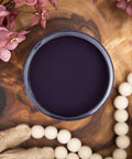 Dixie Belle Paint Aubergine Chalk Mineral Paint is the perfect paint for any DIY project! Image shows an open container of Aubergine Chalk Mineral Paint on top of a wooden stump with beads and flowers.