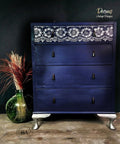 Dixie Belle Paint Black Glaze.
Image shows a dark purple dresser with Black Glaze used on the outer corners fading towards the center to give the dresser dark goth vibes. Top drawer has Lotus Bloom - Stencil in white all along drawer, dresser has black hardware. Staged on a black wall with transparent green glass vase with yellow and red staw .