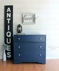 Dixie Belle Paint Bunker Hill Blue Chalk Mineral Paint is the perfect paint for any DIY project!
Dresser painted in Bunker Hill Blue Chalk Mineral Paint with white hardware staged on top of wood floors and walls, Next to piece is a large vertical sign that says "ANTIQUES" and a small white vase with greenery. On top of the piece has a black oil lamp and a book with a white frame that says "FAMILY" hanging above the piece.