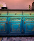 Dixie Belle Paint Bunker Hill Blue Chalk Mineral Paint is the perfect paint for any DIY project!
A credenza painted in a gradient effect with Putty, Limeade, The Gulf, Peacock and lastly the Bunker Hill Blue  all from the Chalk Mineral Paint line as the last darkest color. Staged with two bronze pitchers and a black vintage camera also in front of a pink wall.