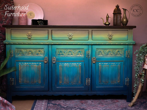 Dixie Belle Paint Bunker Hill Blue Chalk Mineral Paint is the perfect paint for any DIY project!
A credenza painted in a gradient effect with Putty, Limeade, The Gulf, Peacock and lastly the Bunker Hill Blue  all from the Chalk Mineral Paint line as the last darkest color. Staged with two bronze pitchers and a black vintage camera also in front of a pink wall.