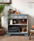 Dixie Belle Paint Buttercream Chalk Mineral Paint is the perfect paint for any DIY project!
A furniture makeover of a small bookcase with drawers on the inside of the bookshelves are painted in Buttercream Chalk Mineral Paint. The Body of the piece is a mute neutral blue. Staged with books, pots and blankets with wooden bowls besides the blanket inside the piece.