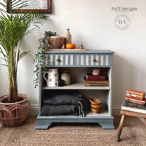 Dixie Belle Paint Buttercream Chalk Mineral Paint is the perfect paint for any DIY project!
A furniture makeover of a small bookcase with drawers on the inside of the bookshelves are painted in Buttercream Chalk Mineral Paint. The Body of the piece is a mute neutral blue. Staged with books, pots and blankets with wooden bowls besides the blanket inside the piece.