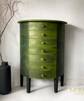 Dixie Belle Paint Copper Bronze Glaze 
This is a round, tall dresser with 7 drawers.  The furniture has 4 legs painted black and keep the dresser approximately 8 inches off the white floor. The entire dresser has had the Copper Bronze Glaze applied to it's surface.  It gives the piece a beautiful sheen.  To the left of the dresser is a large black vase and a leafless branch comes out of the top which ends up being taller than the dresser.  The dresser is in a completely white corner of a room.