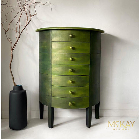 Dixie Belle Paint Copper Bronze Glaze 
This is a round, tall dresser with 7 drawers.  The furniture has 4 legs painted black and keep the dresser approximately 8 inches off the white floor. The entire dresser has had the Copper Bronze Glaze applied to it's surface.  It gives the piece a beautiful sheen.  To the left of the dresser is a large black vase and a leafless branch comes out of the top which ends up being taller than the dresser.  The dresser is in a completely white corner of a room.