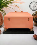 Coral chalk painted jewelry box with gold feet, hinges, and handle. Staged on a black base.