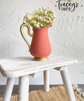 Coral chalk painted pitcher with a gold base and handle. Staged with white flowers inside, on a white painted distressed stool.