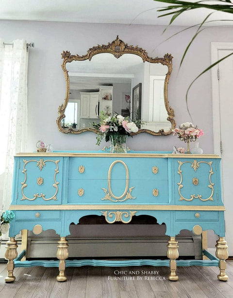 Dixie Belle Paint Dixie Belle Blue Chalk Mineral Paint is the perfect paint for any DIY project!
This is a French, Victorian Style Dresser.  It is painted with Dixie Belle Blue CMP.   There are appliques on both ends and the center of the piece.  The legs are painted in gold just like the appliques and knobs.  There is a gold, ornate mirror on the wall above the dresser and on top it is staged with flowers.