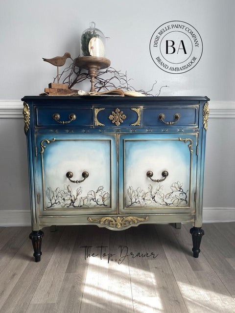 Dixie Belle Paint Dried Sage Chalk Mineral Paint is the perfect paint for any DIY project!
A beautiful buffet with a reverse ombre effect with a blend of blues on top fading down to Dried Sage Chalk Mineral Paint in the center door panels they're white with Bud & Branches - Transfer. Staged with bronze antique items on top with a white wall background.
