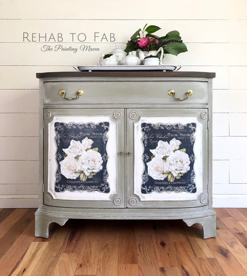 Dixie Belle Paint Dried Sage Chalk Mineral Paint is the perfect paint for any DIY project!
Image shows a bar painted in Dried Sage Chalk Mineral Paint with a painting of a flower on a blue background on door panels. Staged with a tray and a teapot set with greenery behind on top of piece.