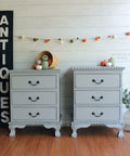 Dixie Belle Paint Driftwood Chalk Mineral Paint is the perfect paint for any DIY project!
A pair of gray chalk painted nightstands staged with fall décor. A large slim black sign with "antiques" written in white letters, faux pumpkins and squash strown on a line above the nightstands, a basket full of faux pumpkins and squash overflowing onto the top of one of the nightstands, and a vase full of faux eucalyptus.