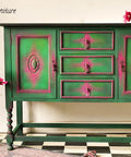 Dixie Belle Paint Evergreen Chalk Mineral Paint is the perfect paint for any DIY project! Green and pink painted dresser with pink flowers placed on top. Glass vase placed on the ground next to the piece containing the same pink flowers. Styled on top of a black and white checkered floor.