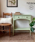 Dixie Belle Paint Farmhouse Green Chalk Mineral Paint is the perfect paint for any DIY project!
Console table painted in Farmhouse Green Chalk Mineral Paint with white hardware, Staged with a wooden chair and a pillow with fringe on it, and books are staged on top of the piece.