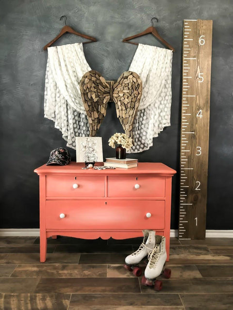 Dixie Belle Paint Flamingo Chalk Mineral Paint is the perfect paint for any DIY project!
Dresser painted in  Flamingo Chalk Mineral Paint with white hardware, staged with angel wings and two curtains meeting at the angel wings hanging on the wall behind the piece. Along with a pair of roller skates in front of the piece with a large ruler plank leaning on the wall.