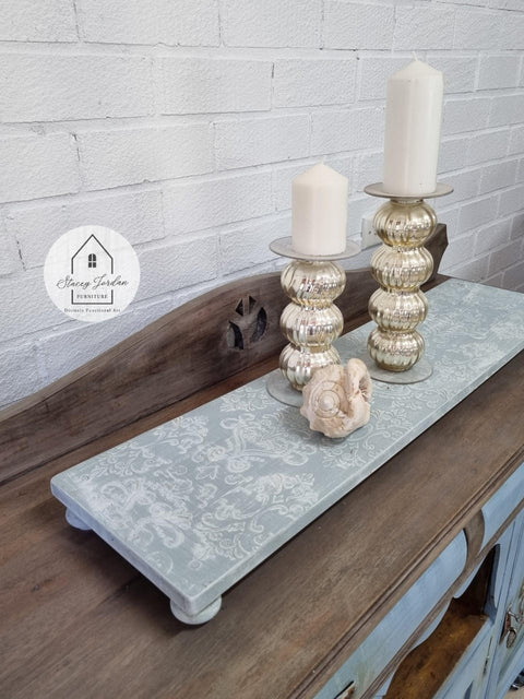 Dixie Belle Paint Haint Blue Chalk Mineral Paint is the perfect paint for any DIY project!
A wooden tray with legs painted with Blue Chalk Mineral Paint  and the Victorian Damask - Stencil in white is seen on the top side of the tray. Staged with two gold candle pedestals with white candles along with a seashell.