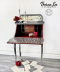 Dixie Belle Paint Hurricane Gray Chalk Mineral Paint is the perfect paint for any DIY project!
Secretary desk staged open, painted in Hurricane Gray Chalk Mineral Paint with the inside the desk is also painted in Muscadine Wine Chalk Mineral Paint and on the inside of the open desk door is decoupaged with Art Deco Bird Pattern - Rice Paper. Staged with ripped pages and red flowers,  as well  as two long silver candle holder placed on top of the piece.