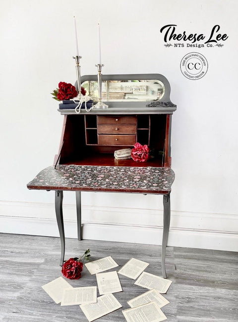Dixie Belle Paint Hurricane Gray Chalk Mineral Paint is the perfect paint for any DIY project!
Secretary desk staged open, painted in Hurricane Gray Chalk Mineral Paint with the inside the desk is also painted in Muscadine Wine Chalk Mineral Paint and on the inside of the open desk door is decoupaged with Art Deco Bird Pattern - Rice Paper. Staged with ripped pages and red flowers,  as well  as two long silver candle holder placed on top of the piece.