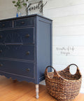 Dixie Belle Paint In the Navy Chalk Mineral Paint is the perfect paint for any DIY project!
A Dresser painted in In the Navy Chalk Mineral Paint  with distressed edges around the dresser. Staged with a woven basket besides the piece along a white wall.