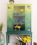 Dixie Belle Paint Kudzu Chalk Mineral Paint is the perfect paint for any DIY project!
China Cabinet painted with from top to bottom in a blended fashion,  Limeade, Kudzu and lastly Palmetto Green Chalk Mineral Paint. Staged with small vases with yellow flowers inside piece. As well as a black woven basket with sticks, yellow and pink flowers with a pruning shears and a pair of working boots to the side of the piece.
